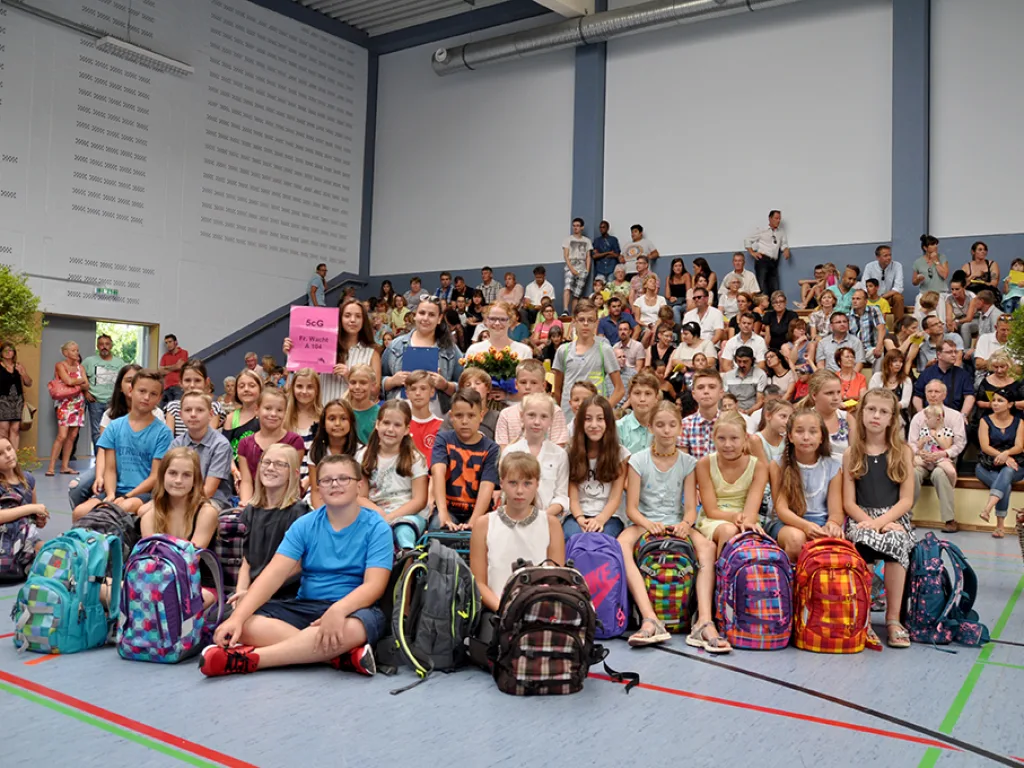 Kinder sitzen mit ihren Ranzen in Reihen auf dem Hallenboden.